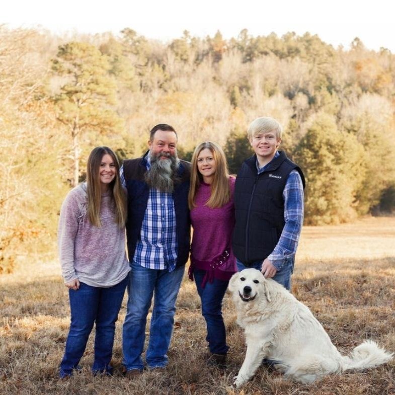 Family portrait featuring four members standing outdoors in a field with a golden retriever. The backdrop showcases a wooded area with fall foliage. The group is dressed casually, smiling, and appears to be enjoying a sunny day together.